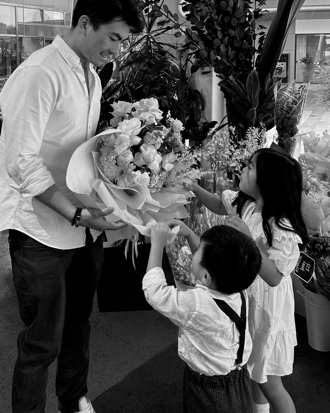 A man in a white shirt holds a bouquet of flowers as two children reach for them in a monochrome photo.