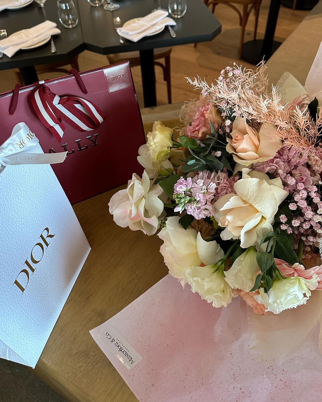Close-up studio shot of a floral bouquet with roses and smaller flowers, wrapped in pink paper, with branded shopping bags.