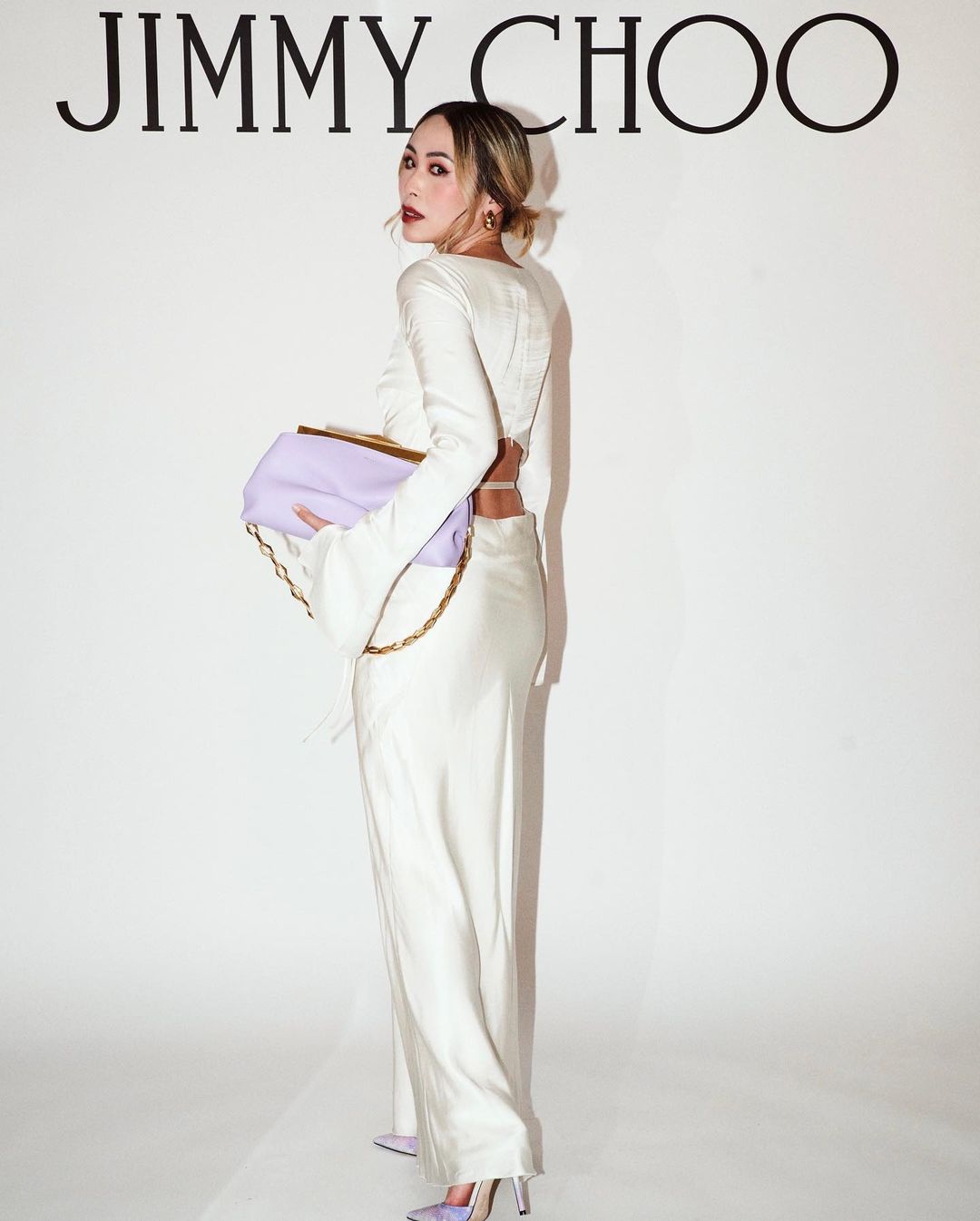 A woman in a white outfit holds a lilac purse with a gold chain in front of a backdrop with the Jimmy Choo logo.