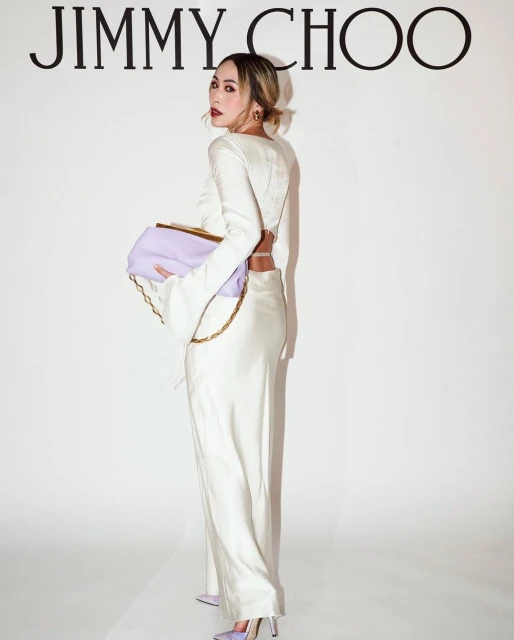 A woman in a white outfit holds a lilac purse with a gold chain in front of a backdrop with the Jimmy Choo logo.
