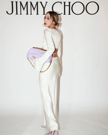 A woman in a white outfit holds a lilac purse with a gold chain in front of a backdrop with the Jimmy Choo logo.