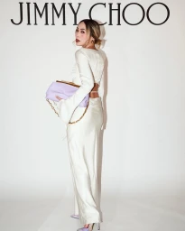 Woman in a white dress holding a lavender shoulder bag standing against a seamless white wall with Jimmy Choo branding.