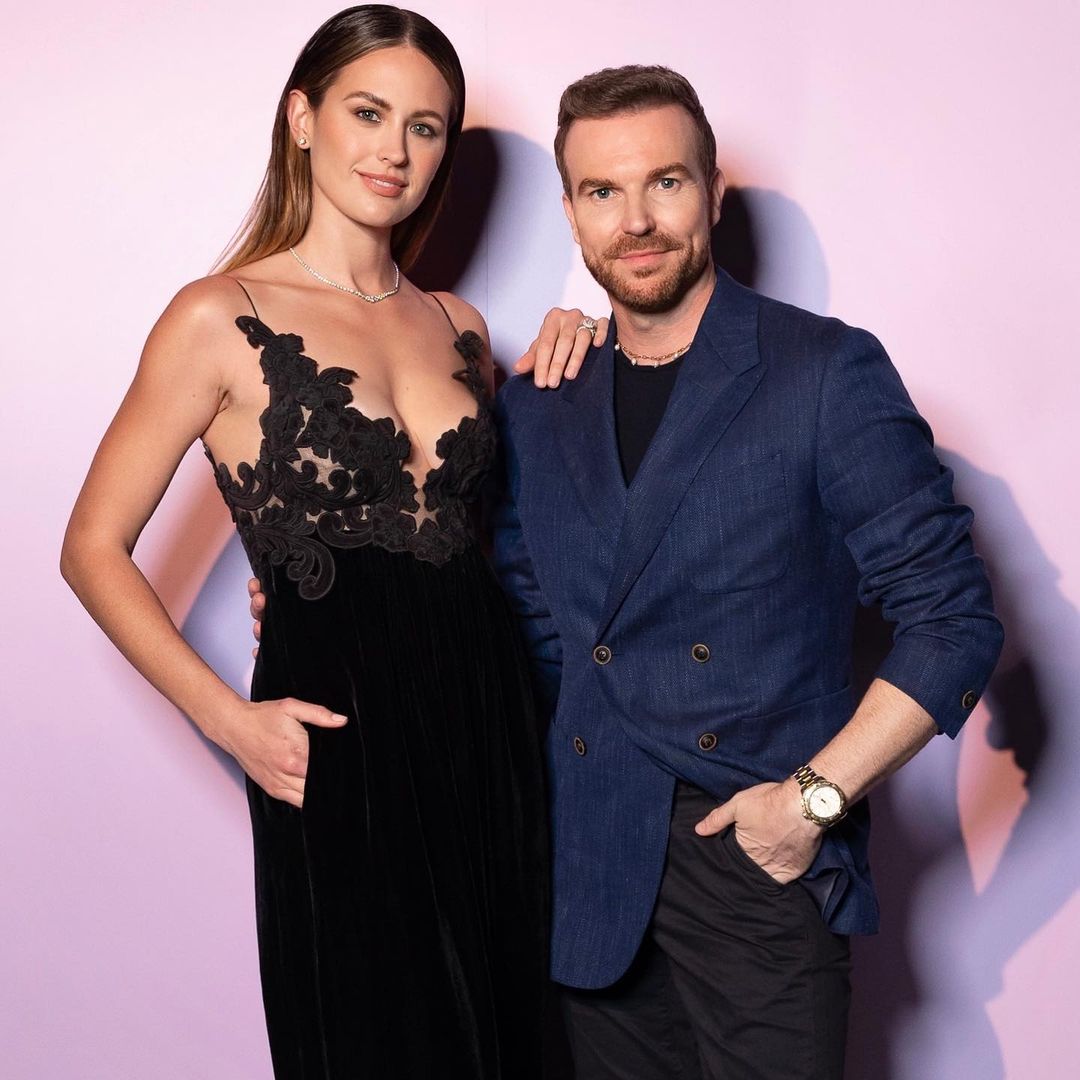 Elegant studio shot of a smiling woman in a black gown with floral lace, posing with a stylish man in a blue suit jacket.
