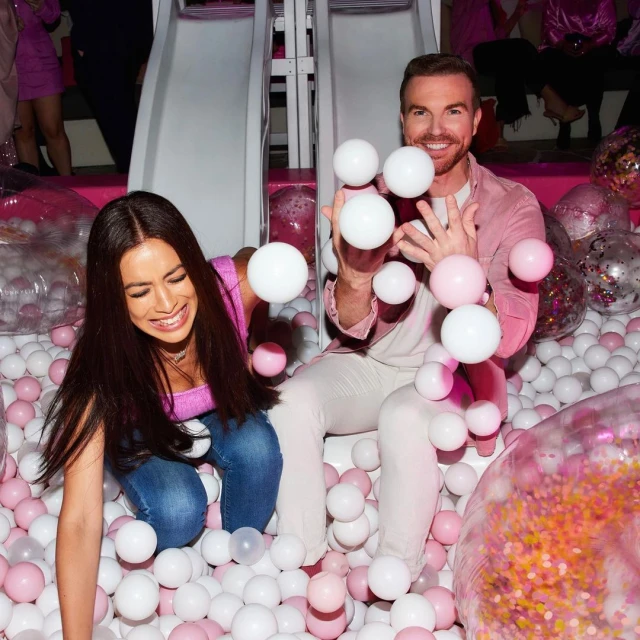 Two adults smiling and throwing pink and white balls in an indoor ball pit with a white slide in the background.