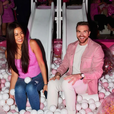 A smiling man and woman sit in a pit filled with white and pink balls, enjoying an event with a white slide in the background.