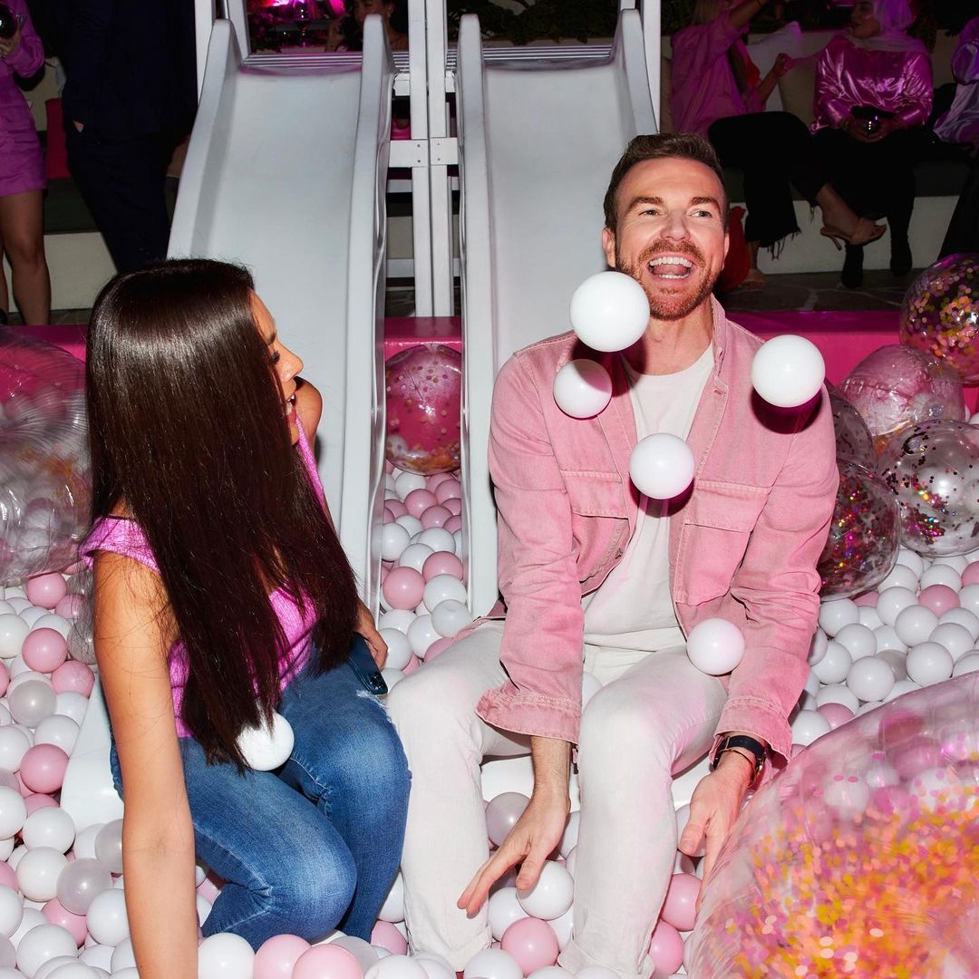 Two people are enjoying a party in a pink and white ball pit with white slides in the background and pink and white confetti balloons.