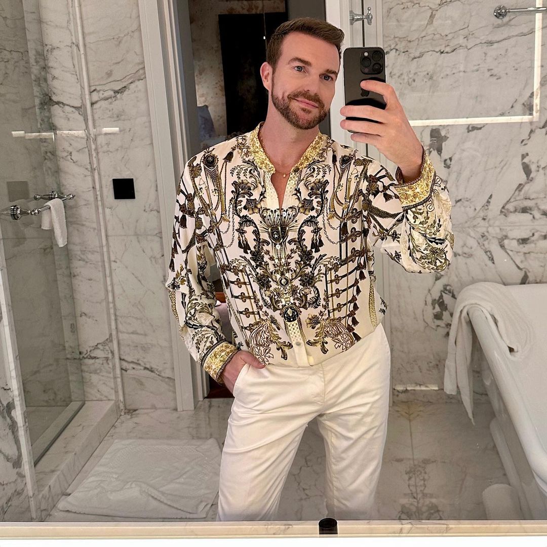 Adult male takes a selfie in a marble bathroom wearing a patterned shirt and white pants, capturing a modern and well-lit interior.