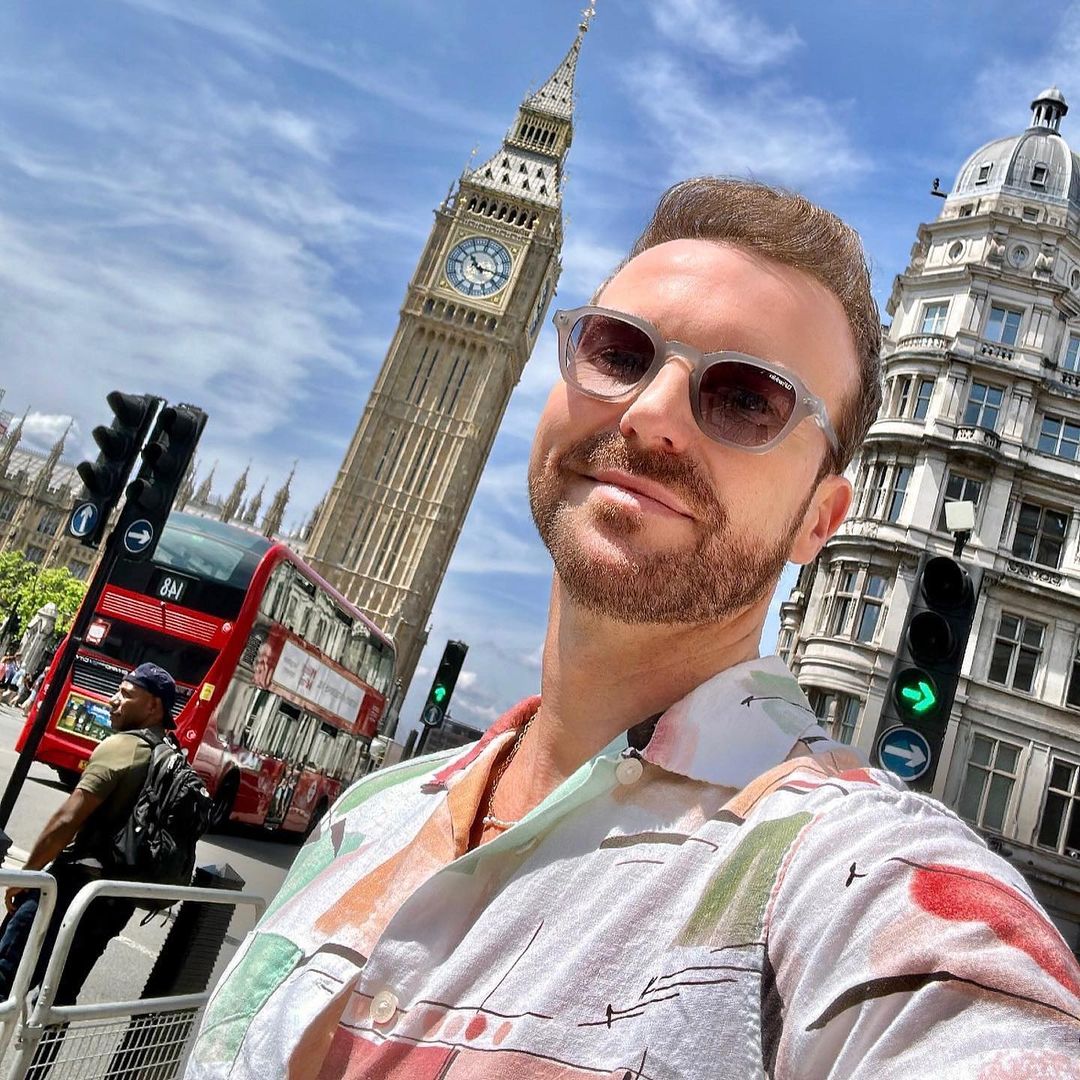 Adult man takes selfie in London, England, with Big Ben, double decker bus, and traffic lights in view on a bright, sunny day.