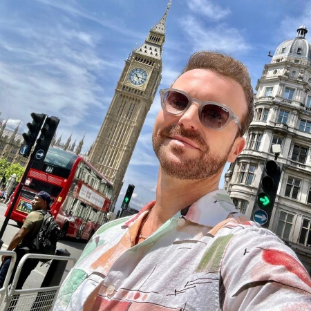 Adult man takes selfie in London, England, with Big Ben, double decker bus, and traffic lights in view on a bright, sunny day.