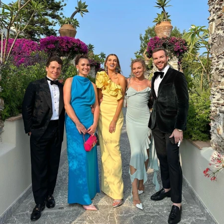 Group of five people in formal wear pose outdoors in a garden setting with bougainvillea, stone walls, and blue sky.