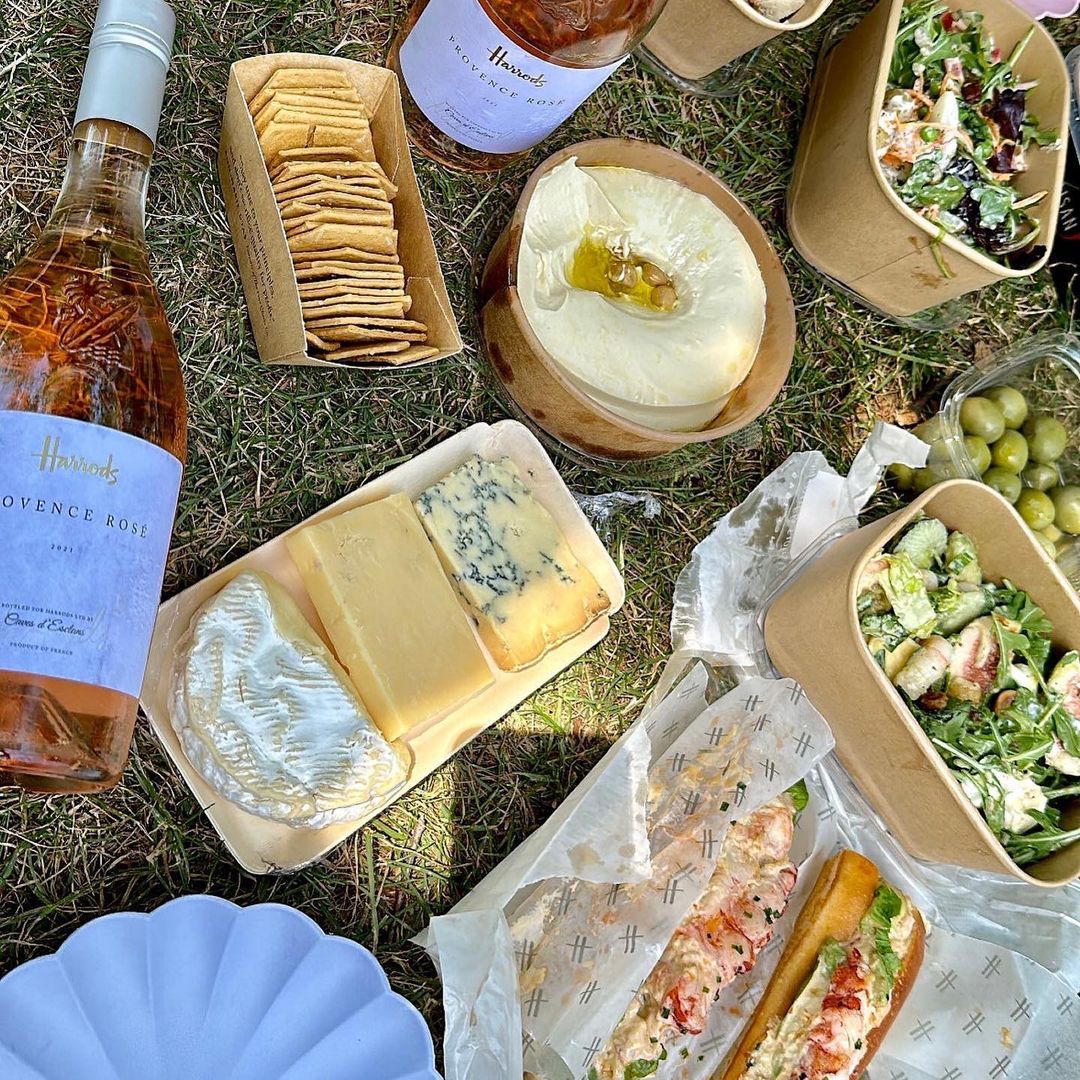 Outdoor picnic featuring wine, cheese, crackers, and salads arranged on a grassy surface, showcasing a selection of food and drink items.