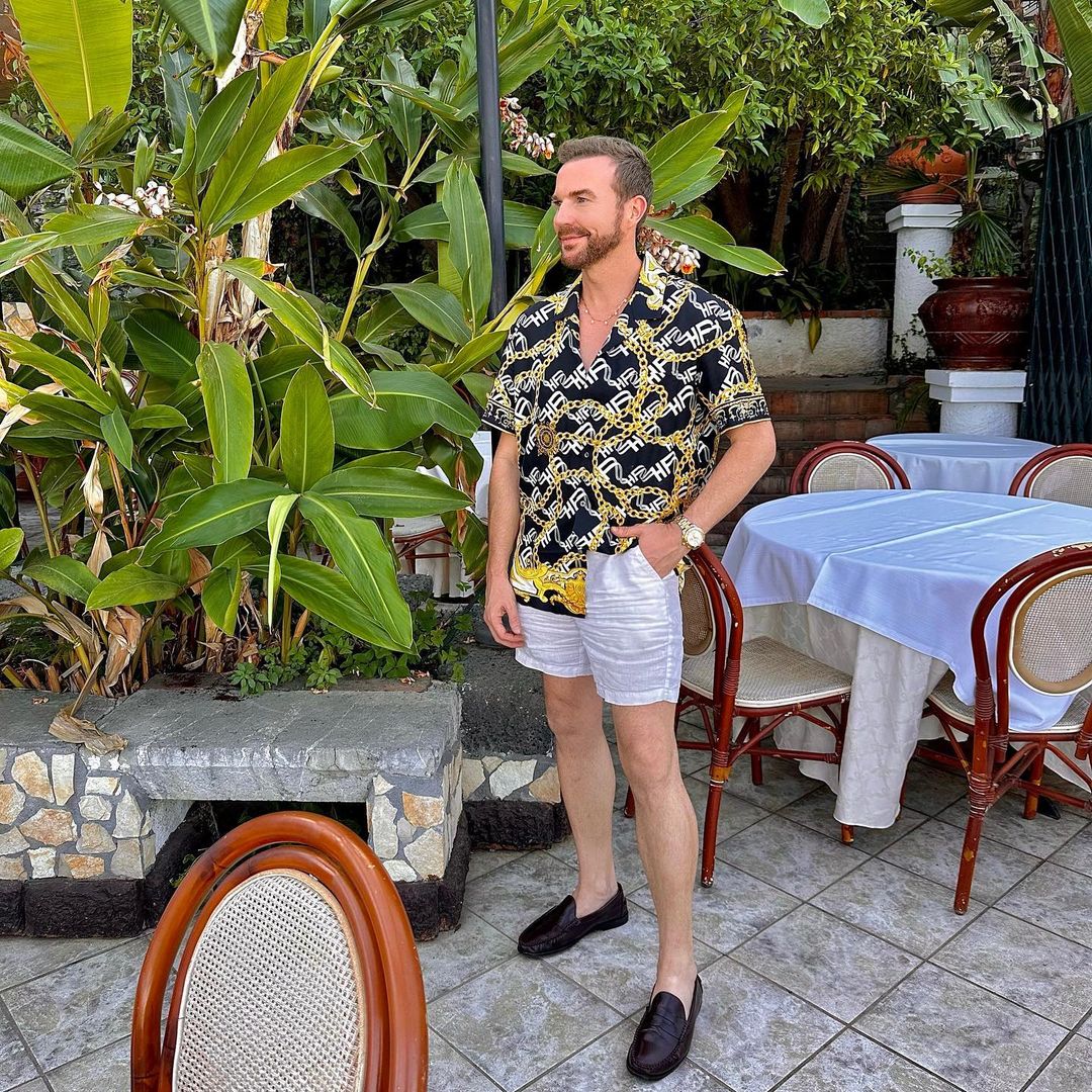 Adult man wearing a patterned shirt and white shorts stands outdoors near plants and tables on a patio, posing for the camera.