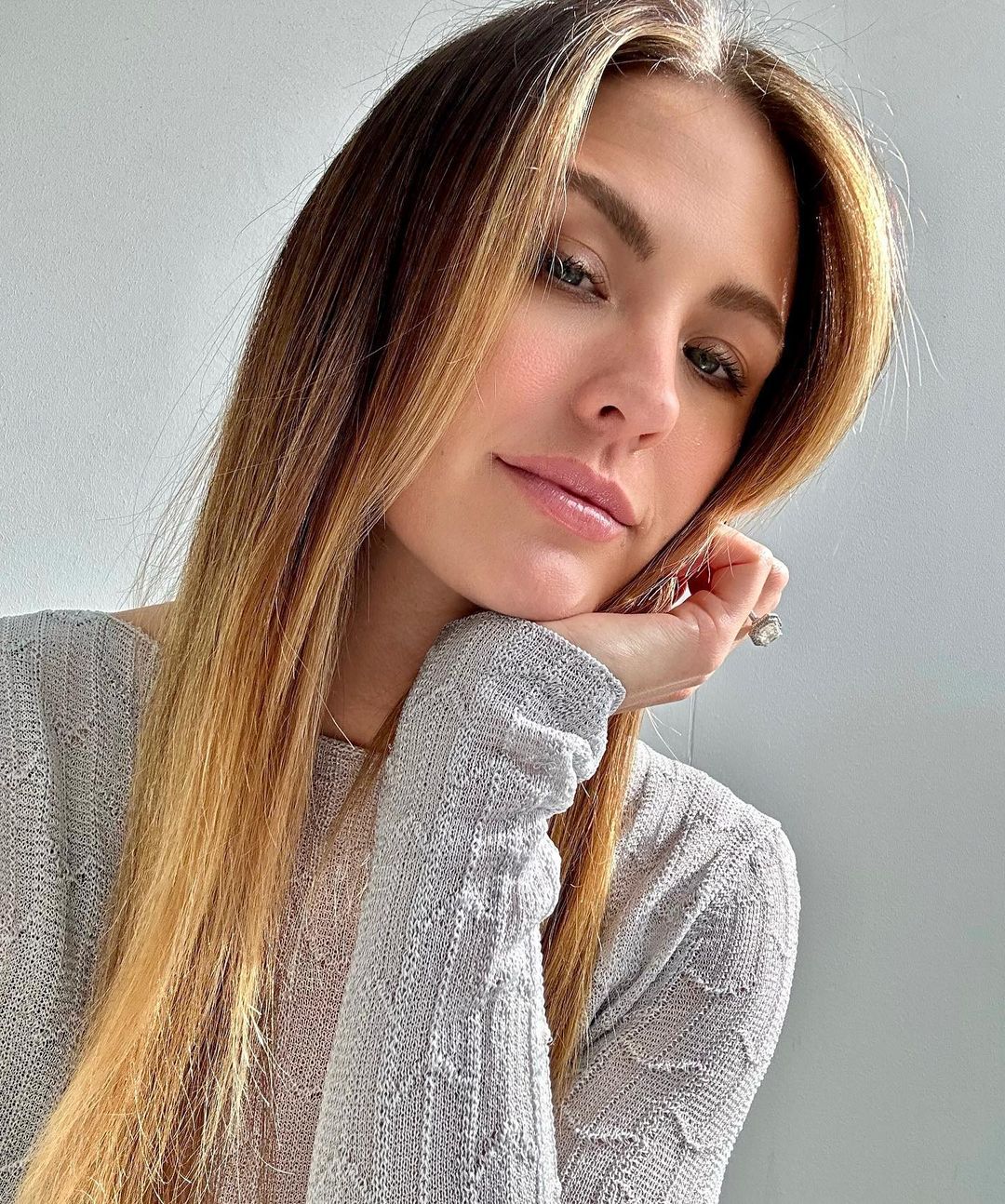 Close-up of a woman with light brown and blonde hair, wearing a light-colored knitted sweater, resting her face on her hand indoors.
