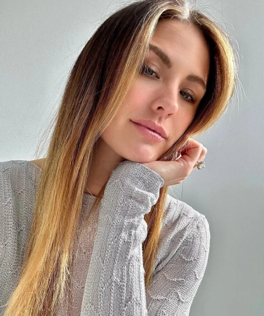 Close-up of a woman with light brown and blonde hair, wearing a light-colored knitted sweater, resting her face on her hand indoors.