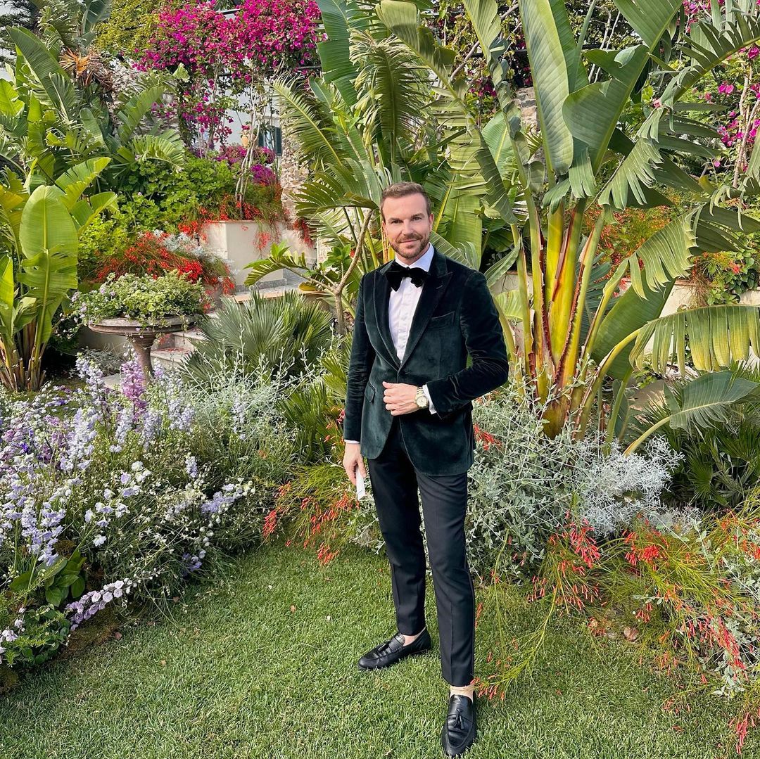 Man in a dark green velvet suit and bow tie stands in a lush green outdoor garden with vibrant flowers and greenery in the background.