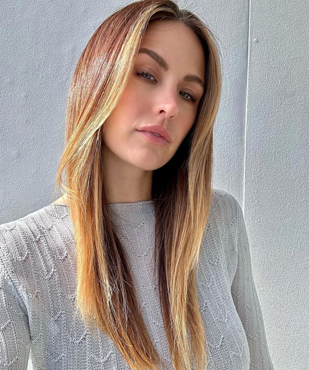 Close-up shot of a light-skinned woman with long, highlighted brown hair and wearing a light gray sweater against a neutral background.