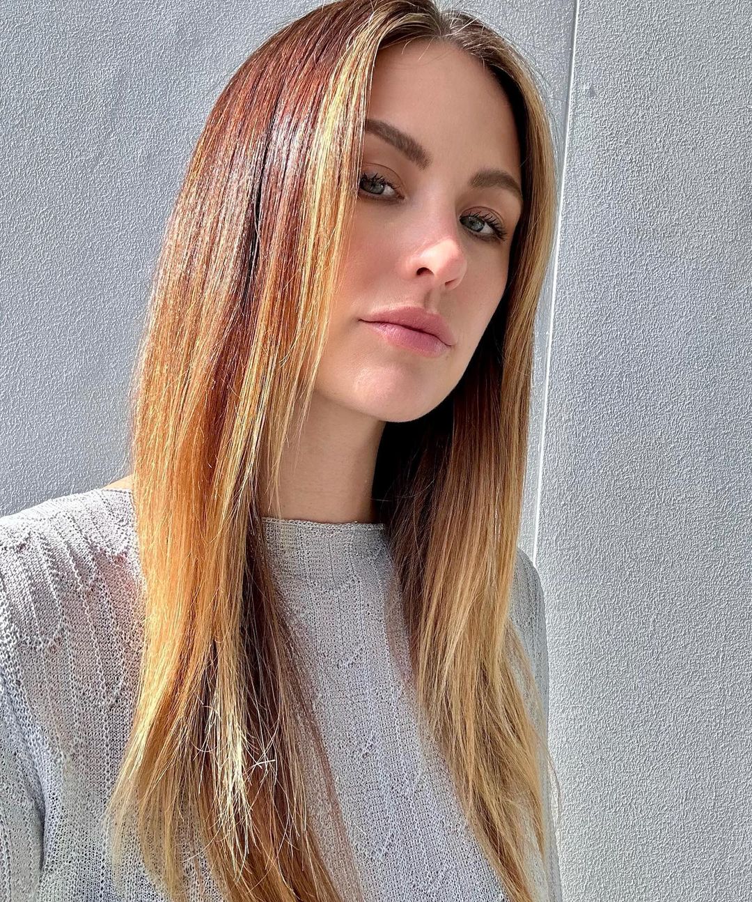 Close-up portrait of an adult woman with long, highlighted brown hair wearing a grey knitted sweater, set against a plain grey background.