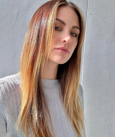 Close-up portrait of an adult woman with long, highlighted brown hair wearing a grey knitted sweater, set against a plain grey background.