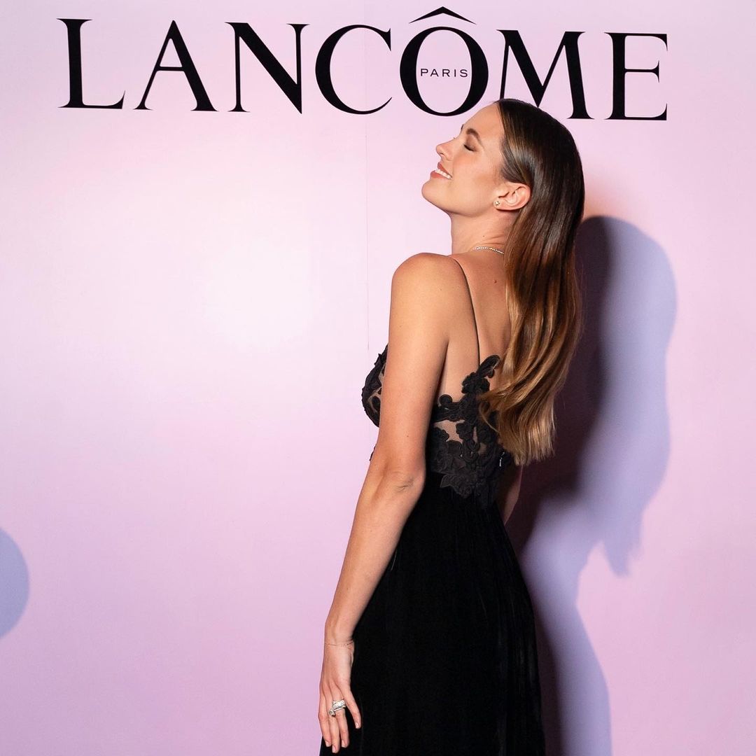 Adult woman in an elegant black dress posing in front of a Lancôme logo on a pink background in an indoor setting. 