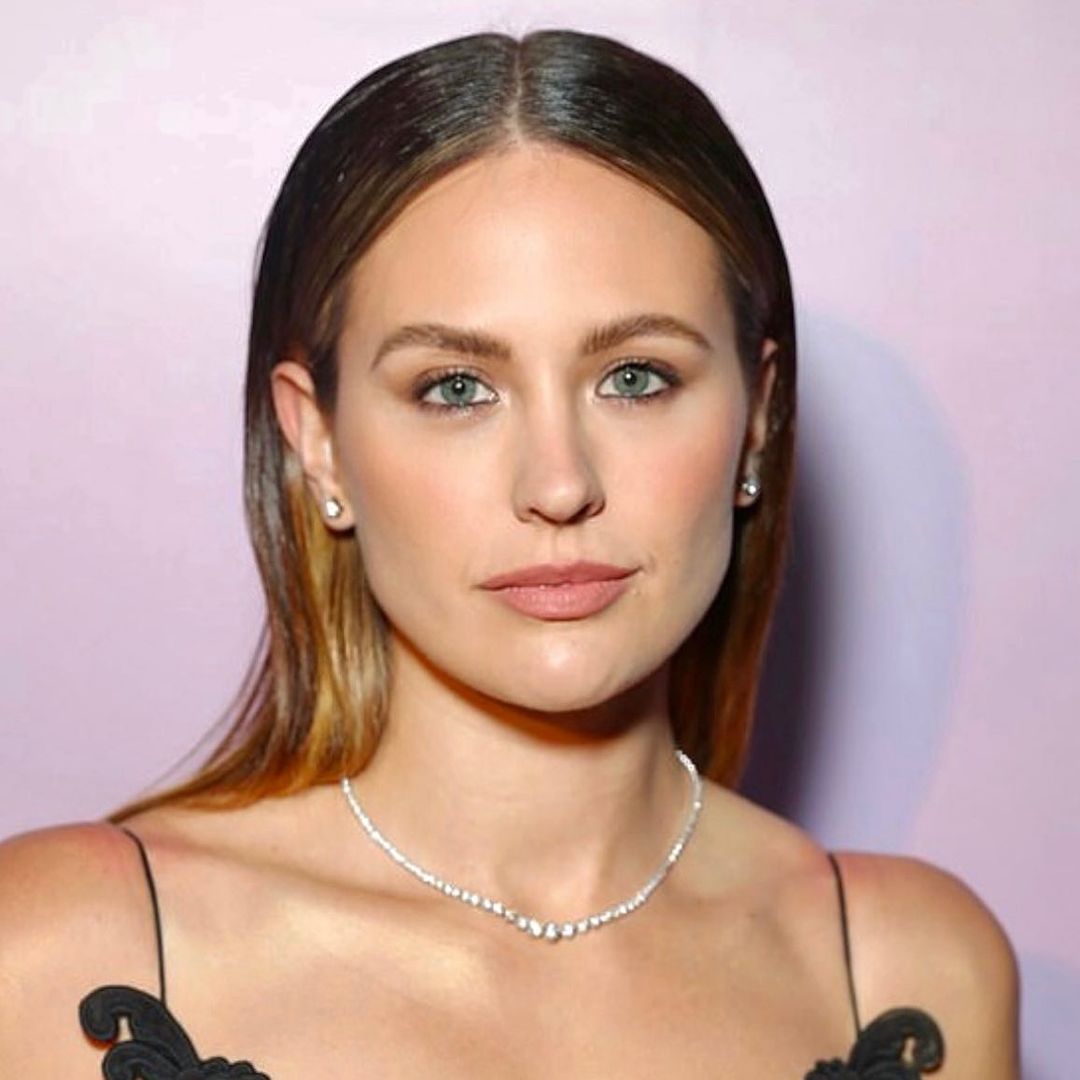 Close-up studio portrait of a stylish woman with light brown hair, fair skin, light eyes, and delicate jewelry posing indoors.