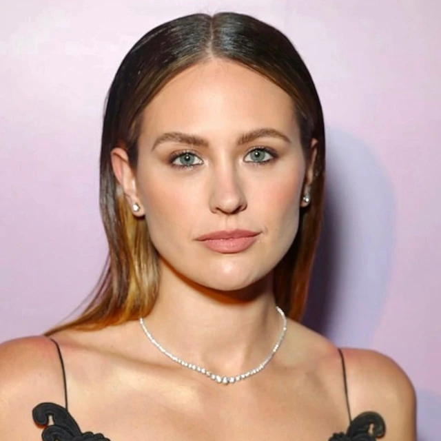 Close-up studio portrait of a stylish woman with light brown hair, fair skin, light eyes, and delicate jewelry posing indoors.