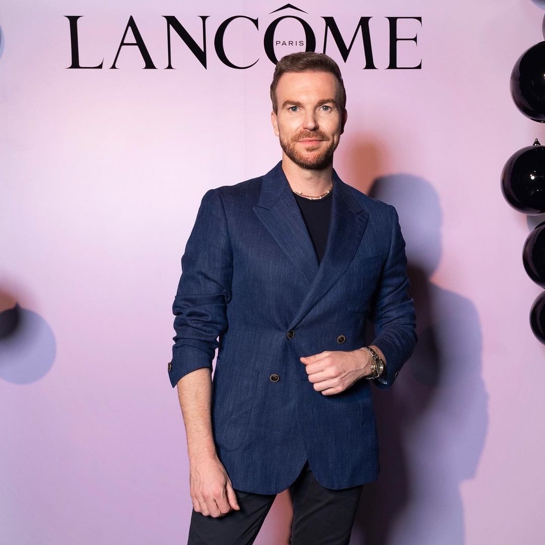 Adult man in a blue blazer stands in front of a Lancome backdrop with black balloons, posing for a portrait in an indoor setting.