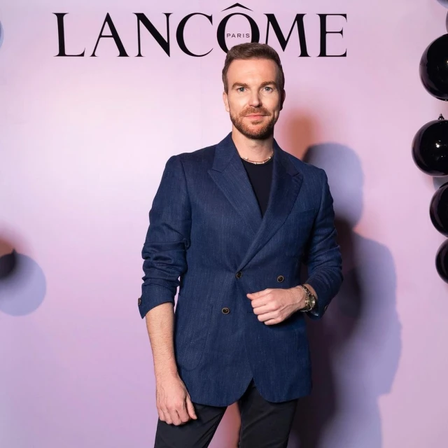 Adult man in a blue blazer stands in front of a Lancome backdrop with black balloons, posing for a portrait in an indoor setting.