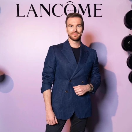Adult man in a blue blazer stands in front of a Lancome backdrop with black balloons, posing for a portrait in an indoor setting.