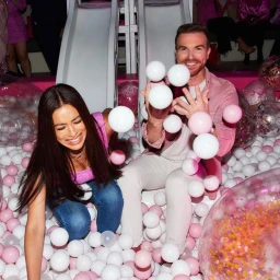 Man and woman smiling in a ball pit filled with white and pink plastic balls, with slides in background