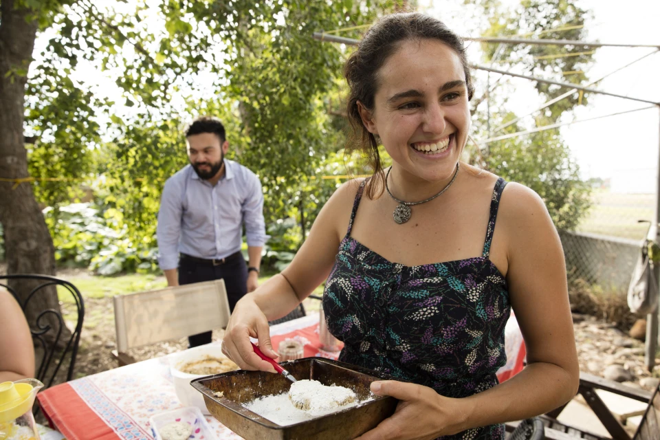 Smiling woman dips food in batter at outdoor table, man stands in background. Sunny day, trees, meal preparation, positive vibe.