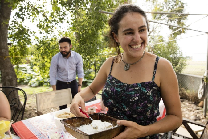 Smiling woman dips food in batter at outdoor table, man stands in background. Sunny day, trees, meal preparation, positive vibe.