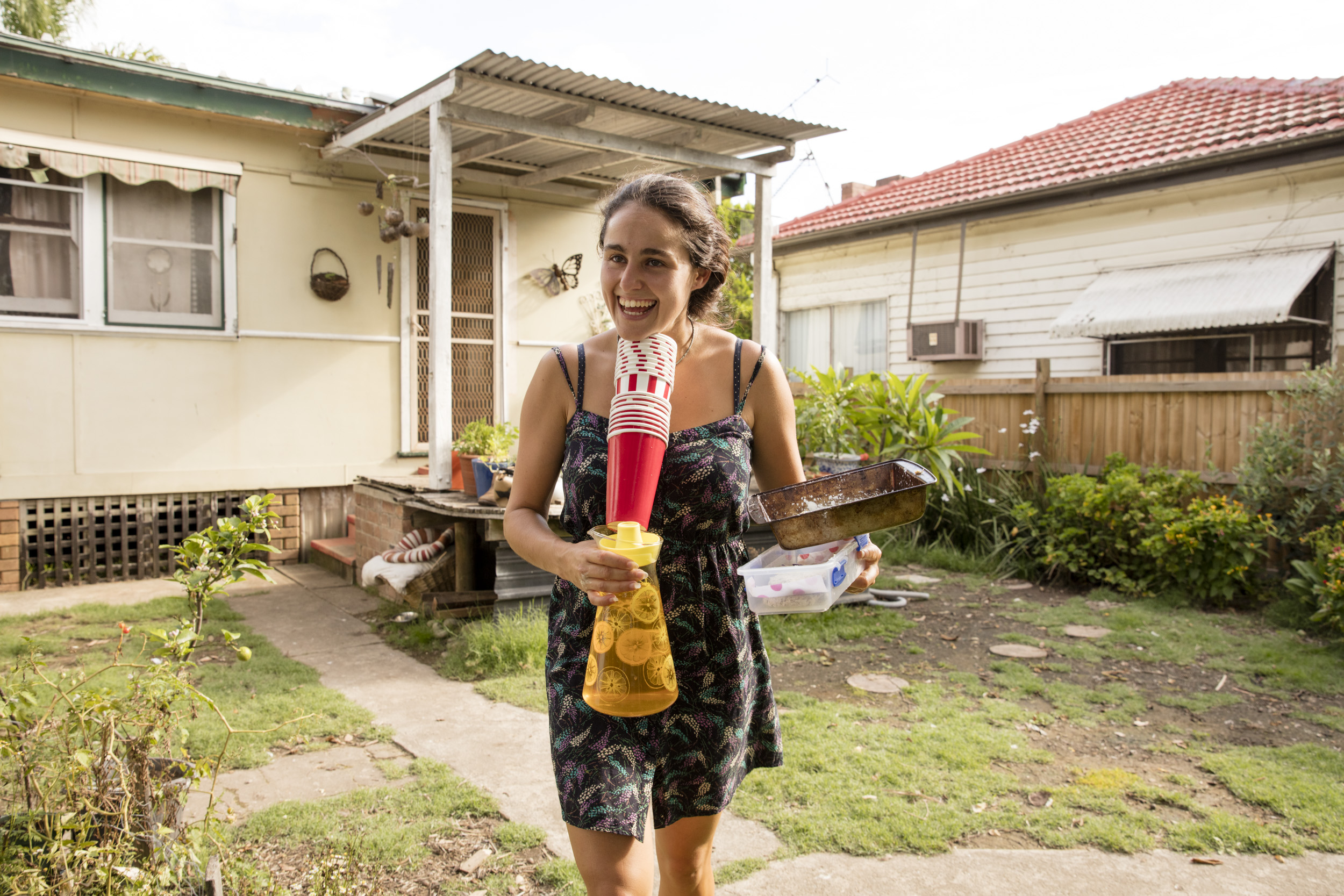 Smiling woman in floral dress stands in yard with baking pan, beverage dispenser with cups, houses, and green grass.