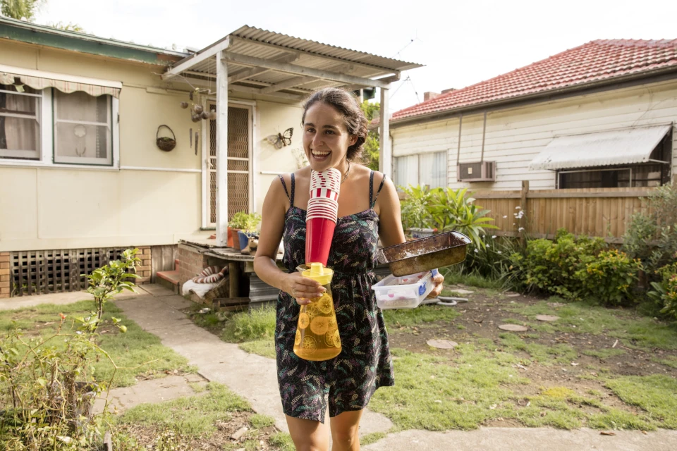 Smiling woman in floral dress stands in yard with baking pan, beverage dispenser with cups, houses, and green grass.
