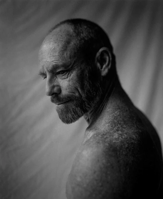 Black and white portrait of a mature man with a beard, gazing off to the side, against a soft, textured backdrop.