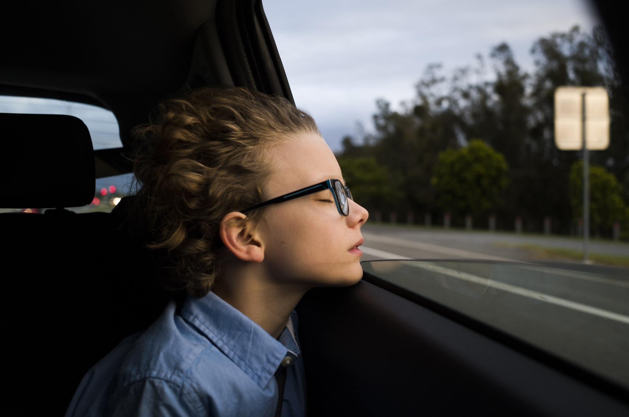 Side view of a child with curly hair, wearing glasses, looking out the car window with trees and a highway visible in the background.