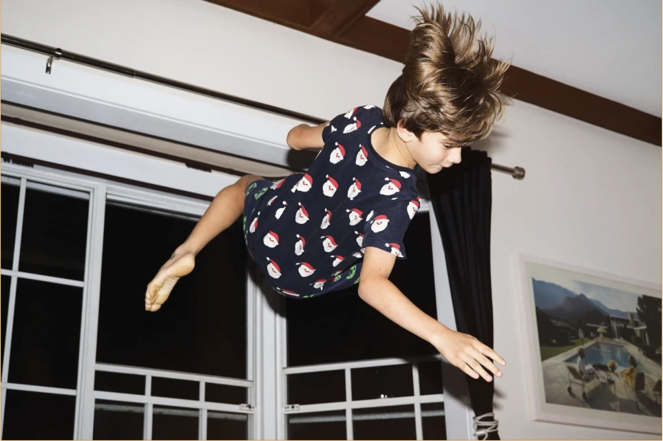 A young boy in mid-air wearing a dark blue shirt patterned with Santa Claus designs, captured indoors near a white framed window.