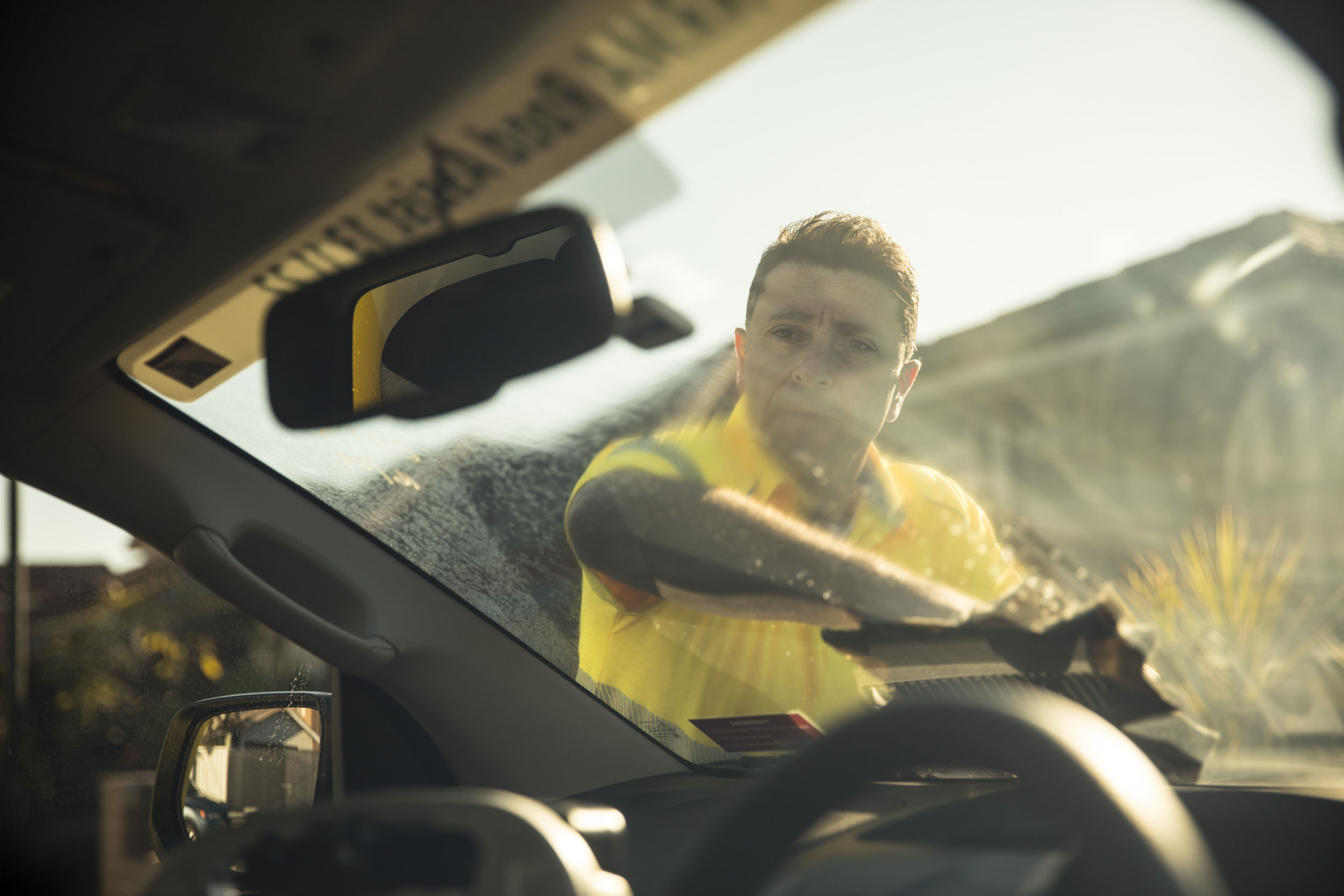 A man in a yellow shirt washes a car windshield from the inside. Light streams in, highlighting the vehicle's interior.