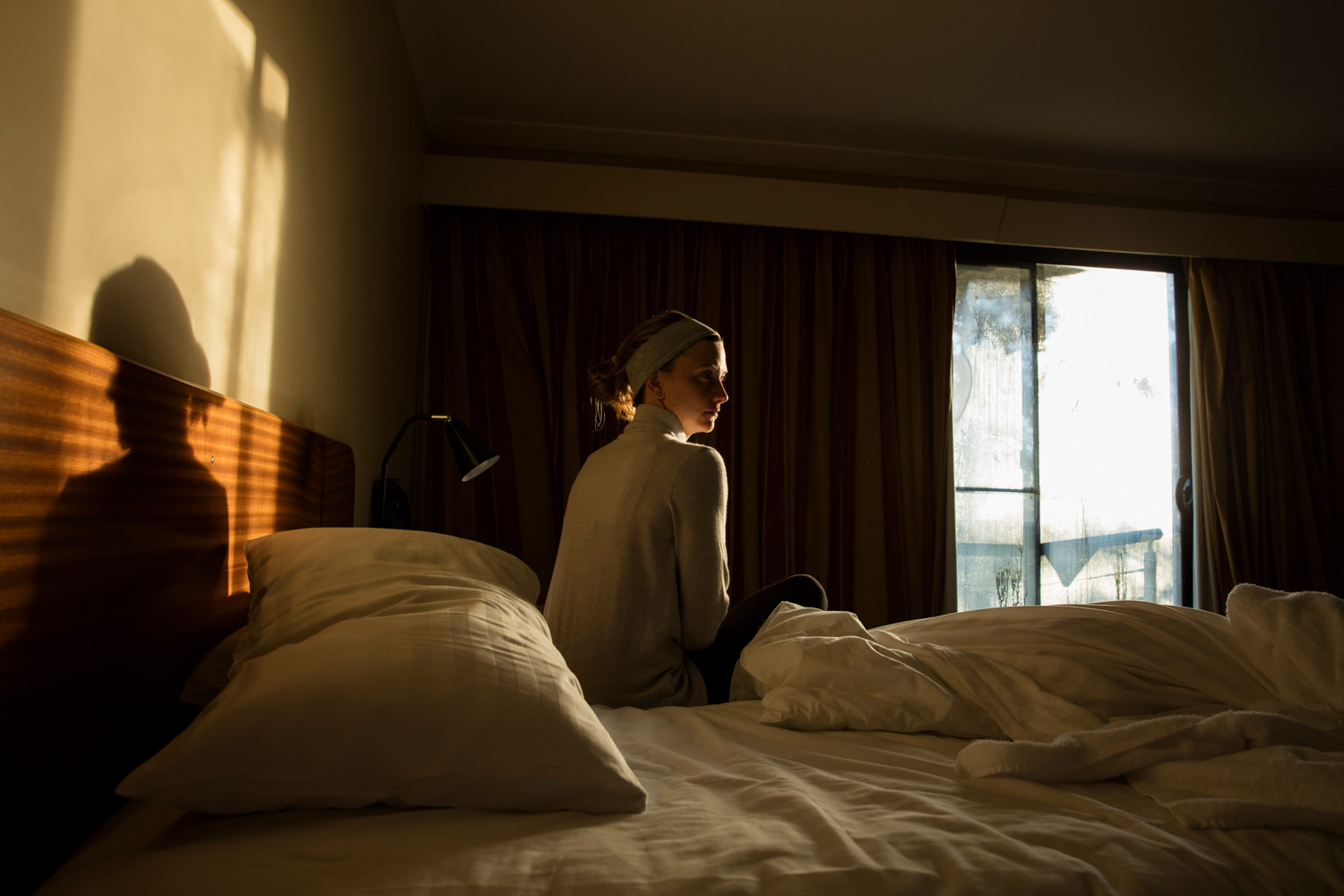 Woman sitting on a bed in a warmly lit room near a window with curtains; the scene is cozy and serene in soft, natural light.