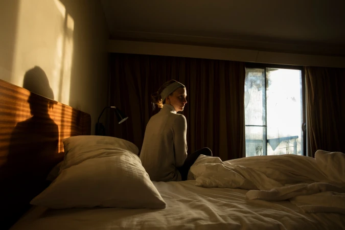 A woman seated on a bed in a hotel room, facing a window with warm morning light and casting a dark shadow on the headboard wall.