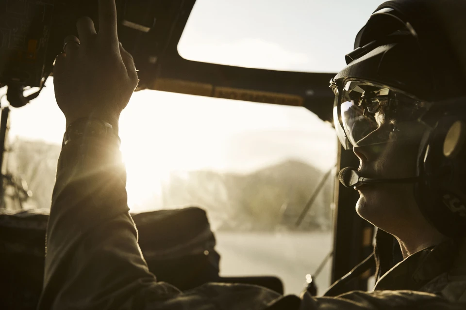 Inside a helicopter cockpit, a pilot in a helmet looks toward the sunlit horizon, with hand near controls in warm, golden lighting.