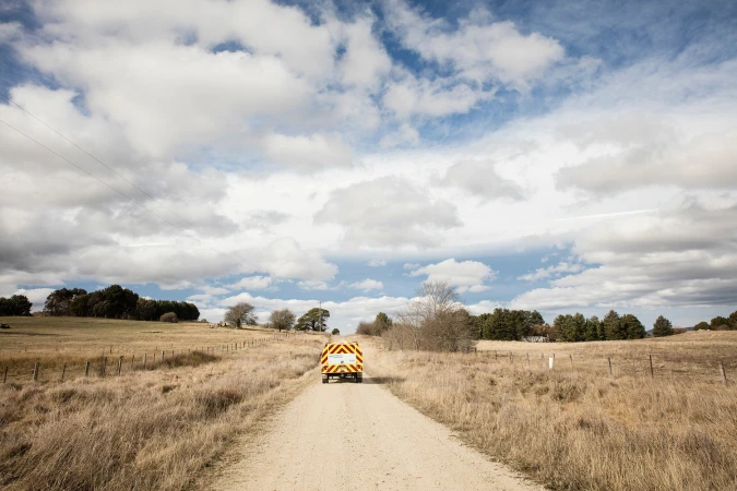 Vehicle drives along dirt road flanked by fields under a cloudy sky, emphasizing rural transport and landscape in a natural setting.