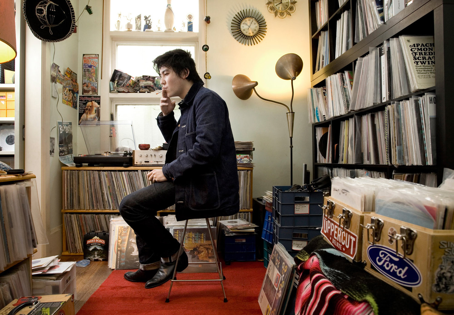 Man smokes indoors sitting on a stool in a room filled with vinyl records, a turntable, a floor lamp, and vintage wall clocks.