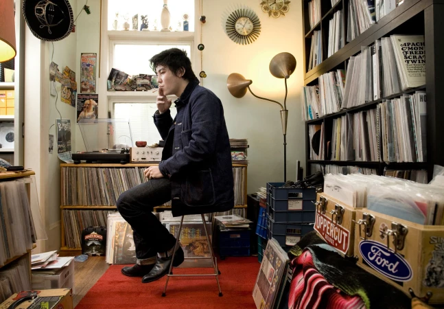 Man smokes indoors sitting on a stool in a room filled with vinyl records, a turntable, a floor lamp, and vintage wall clocks.