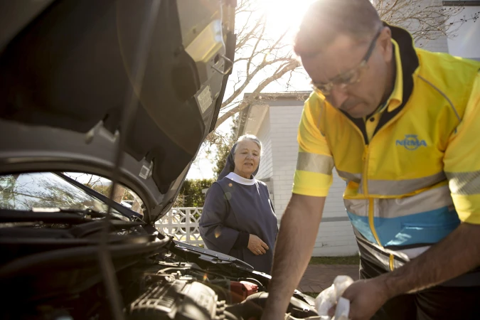 Mechanic in yellow safety vest repairs a car engine with a nun standing nearby outside during the day with a house in the background.