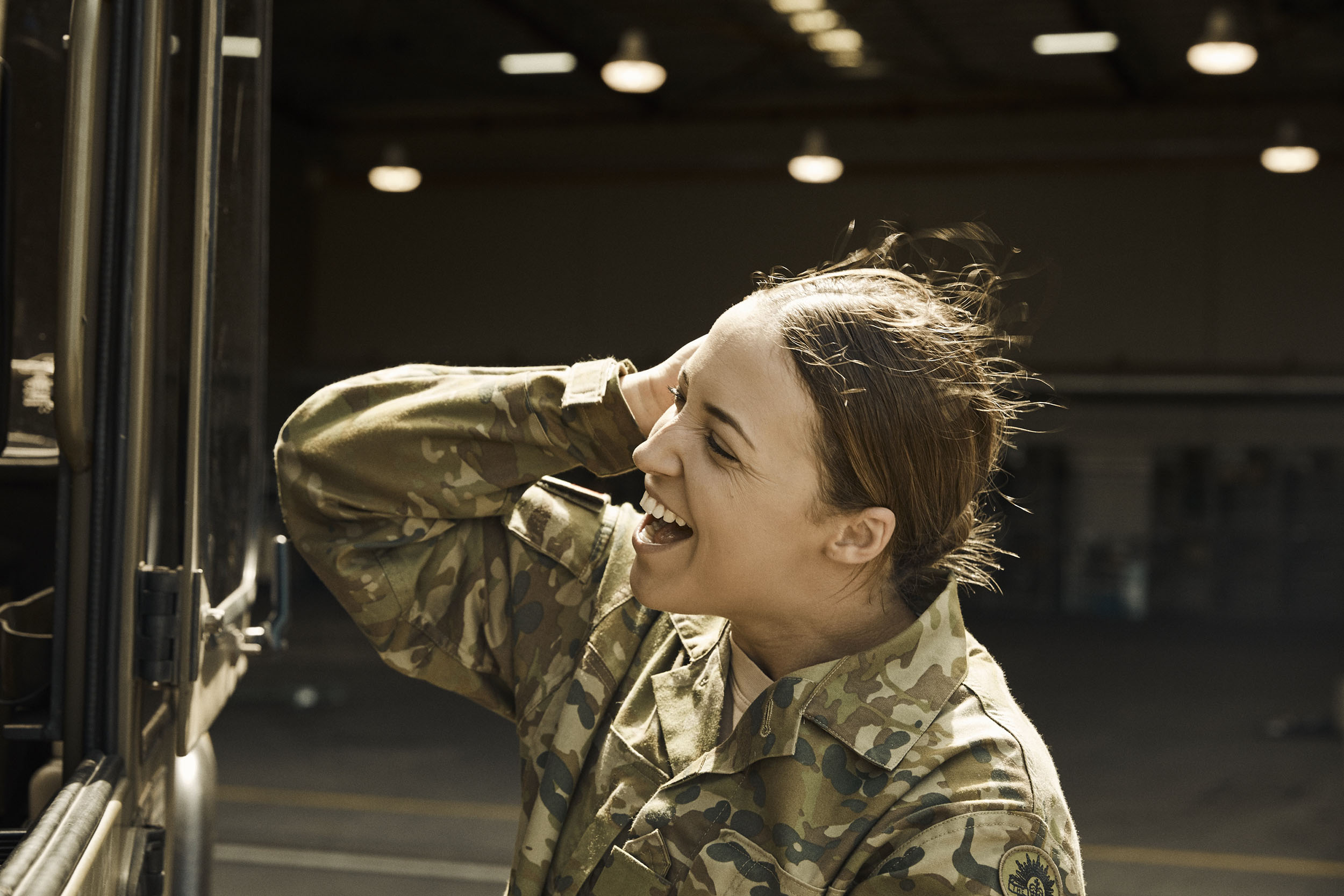 A woman in a camouflage uniform laughs outdoors, with an industrial building visible in the background, during the daytime.