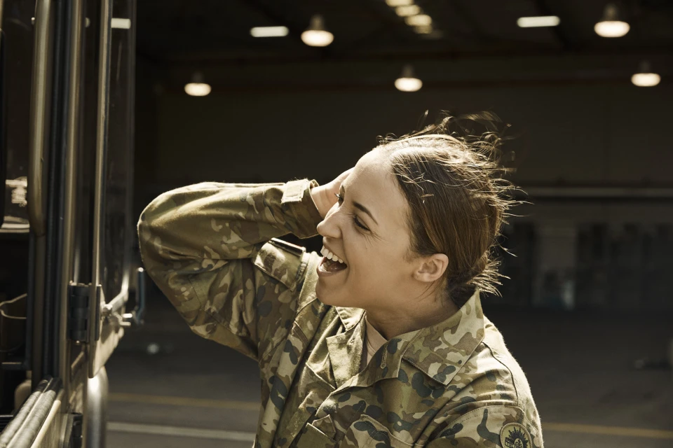 A woman in a camouflage uniform laughs outdoors, with an industrial building visible in the background, during the daytime.