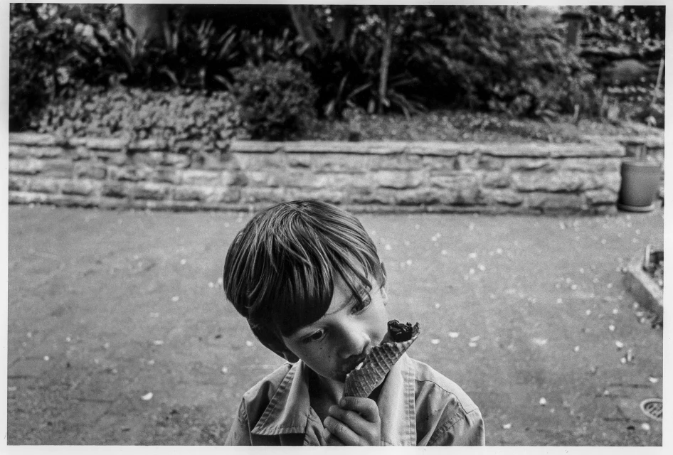 Child enjoys an ice cream cone outdoors with a stone wall and greenery behind in a black and white photograph.