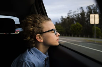 Young person with glasses resting head against a moving car window, captured by Christopher Ireland.