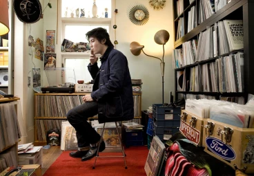 Man seated on a stool smoking a cigarette in a room lined with shelves filled with records, photographed by Christopher Ireland in Sydney.