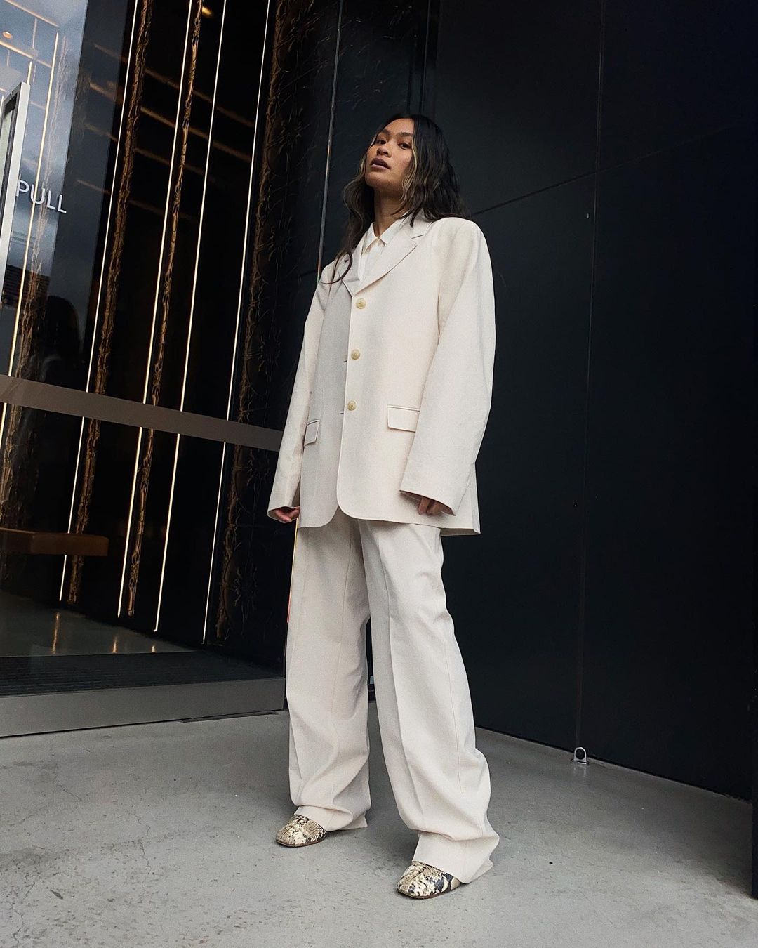 A woman in an oversized off-white suit poses outdoors against a dark, modern building. The outfit is complemented by snakeskin shoes.
